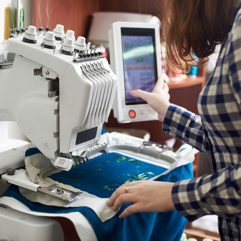 Close-up of woman working on modern computerized embroidery machine creating green floral pattern.
